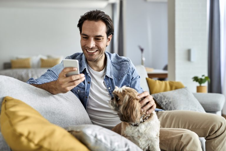 Junger glücklicher Mann schreibt Textnachrichten auf dem Handy, während er mit seinem Hund auf dem Sofa sitzt.