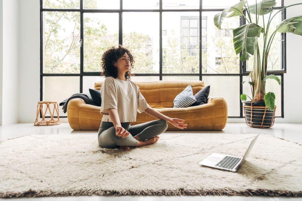 Junge, gemischtrassige Latina-Frau meditiert zu Hause mit Online-Video-Meditationsstunde am Laptop. Meditations- und Spiritualitätskonzept.