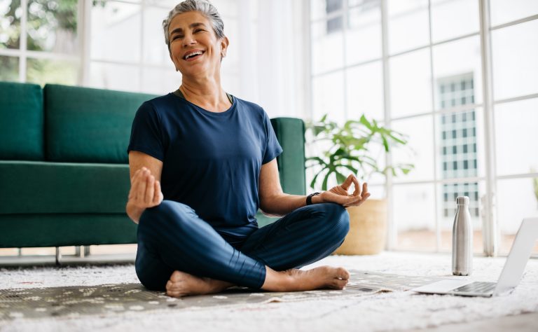 Lächelnde ältere Frau meditiert zu Hause, sie sitzt im Lotussitz vor einem Laptop und verfolgt einen virtuellen Yoga-Kurs. Reife Frau praktiziert Yoga für ihr geistiges und körperliches Wohlbefinden.