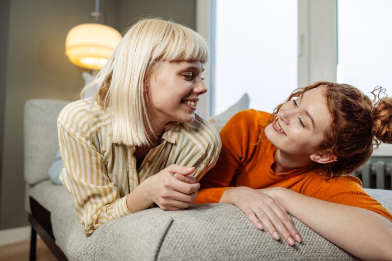 Zwei junge kaukasische Freundinnen liegen auf einem Sofa und lächeln, während sie einander anschauen.