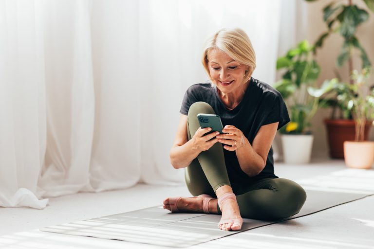 Eine glückliche blonde Seniorin nutzt eine App auf ihrem Mobiltelefon, um Yoga zu machen